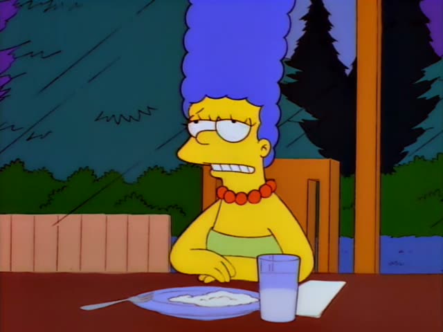Marge sits at a wooden table outdoors in the rain, a plate with a spoonful of food, a fork and a glass of water in front…