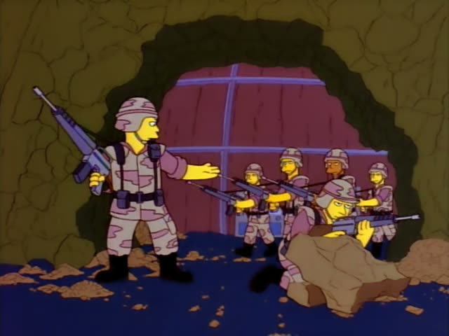 Several one-off soldiers in pink-and-tan camouflage, along with a red-bearded man in a green sweater, stand in a rocky…
