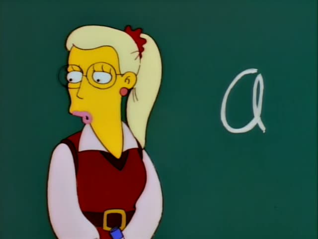 Mrs. Krabappel and a red vest over a white blouse, stands before a green chalkboard with a large chalk letter A and gestures…