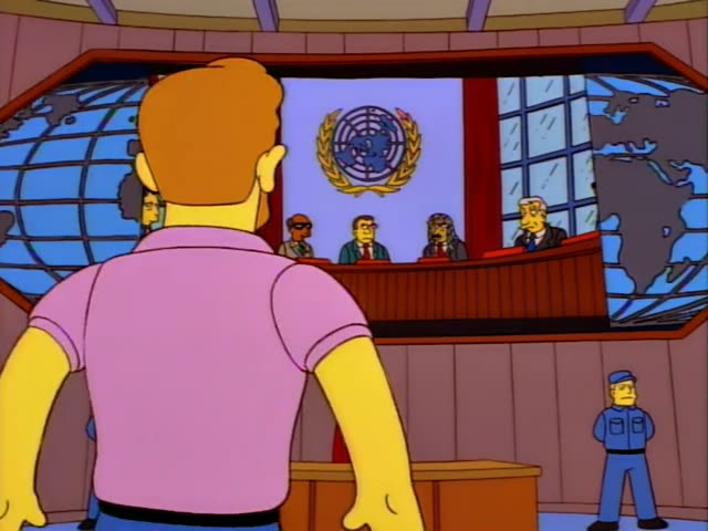 A man with orange hair and a pink polo shirt, seen from behind at first, stands in a UN-style assembly hall with a globe…