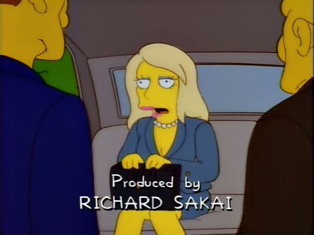 A blonde woman with a pearl necklace and blue blazer sits in the back of a car between two men, one in a blue jacket…