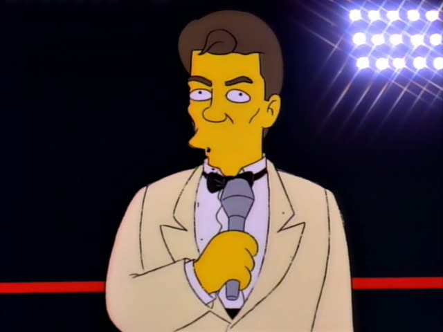 A tuxedo-clad announcer with brown hair and a bow tie holds a microphone and a card in a boxing-ring-style setting lit…