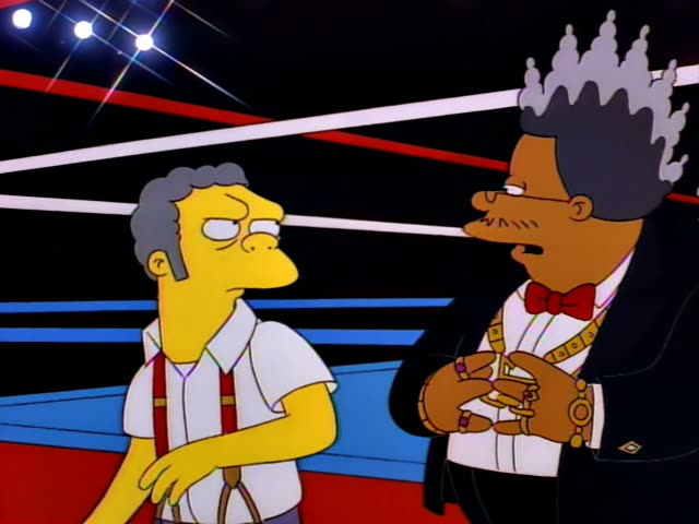 Moe stands glaring in a boxing ring with red and white ropes and stage lights overhead