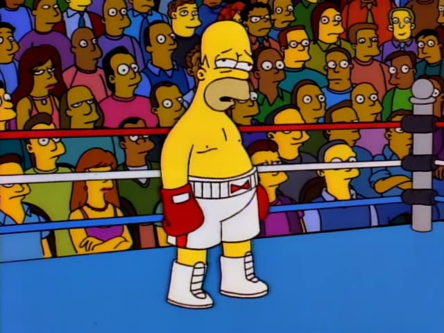 Homer Simpson stands shirtless in white boxing trunks, red gloves and white wrapped boots on a blue ring canvas, surrounded…