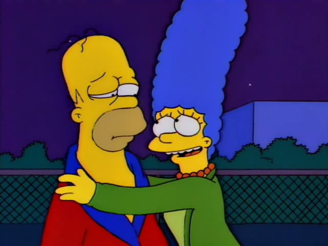 Homer Simpson and Marge Simpson stand together at a chain-link fence at night under a purple sky with bushes and a blue…
