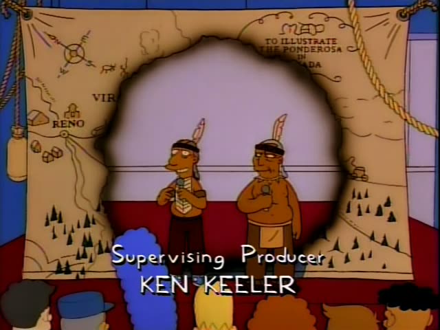 Two men in feathered headbands and fringed costumes stand on a stage in front of a painted map backdrop…