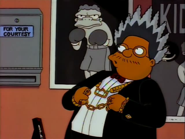 A heavyset man with spiky grey hair, glasses, a mustache, a black suit and gold chains stands in a bar office, first…
