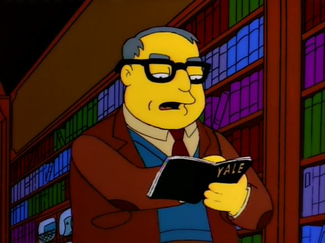 A stout, grey-haired man in glasses, a brown jacket and a red tie stands in a library before tall bookshelves…