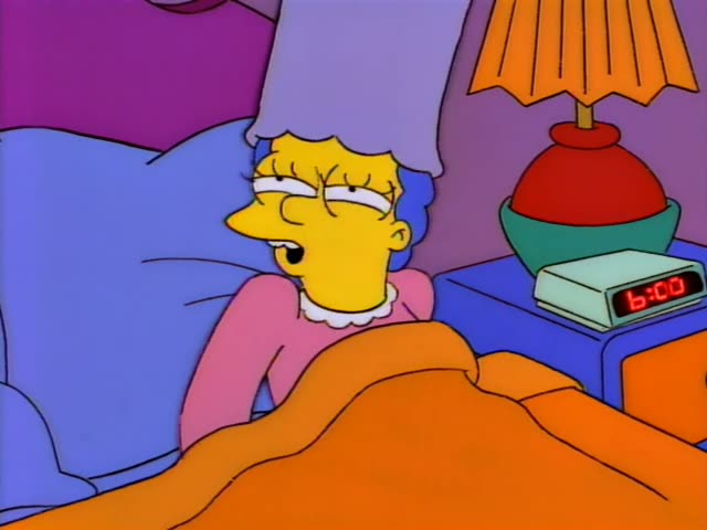 Marge Simpson lies in bed under an orange blanket, pink nightgown and white collar, beside a nightstand with a red-and-green…