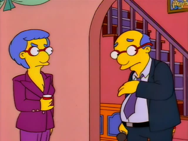 A bald, bespectacled man in a dark suit and purple tie is talking with a woman with short blue hair, red glasses…