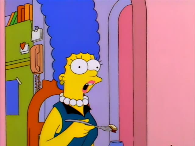 Marge Simpson stands in a kitchen doorway beside a green refrigerator, holding a fork with a bite of food, first smiling…