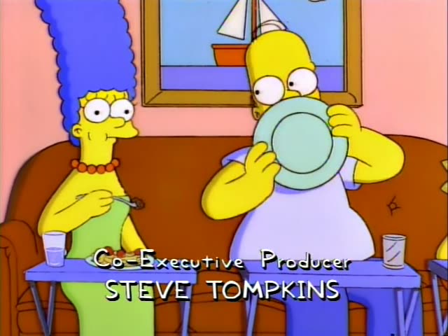 Marge Simpson sits on the brown couch with a fork over a plate of spaghetti while Homer Simpson raises a pale green plate…