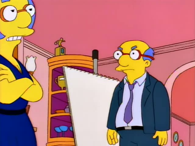 A blue-haired woman in a navy dress with red-framed glasses stands with her arms crossed while a heavyset bald man in a dark…