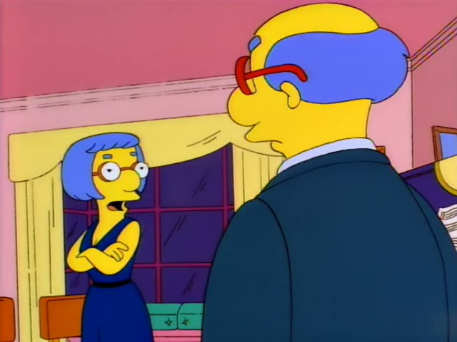 A blue-haired woman in a navy sleeveless dress and red-framed glasses faces a bald man in a dark suit with red glasses, seen…