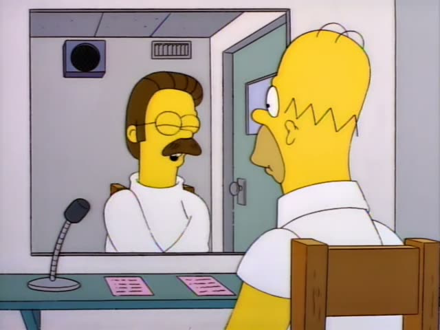 Homer Simpson sits facing a moustached man with glasses in a white shirt who talks to him through a glass partition…
