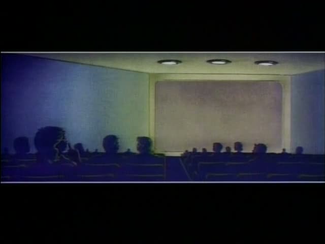 Silhouetted audience members, one with a large round bushy head in the left foreground, sit in rows of seats in a dark…