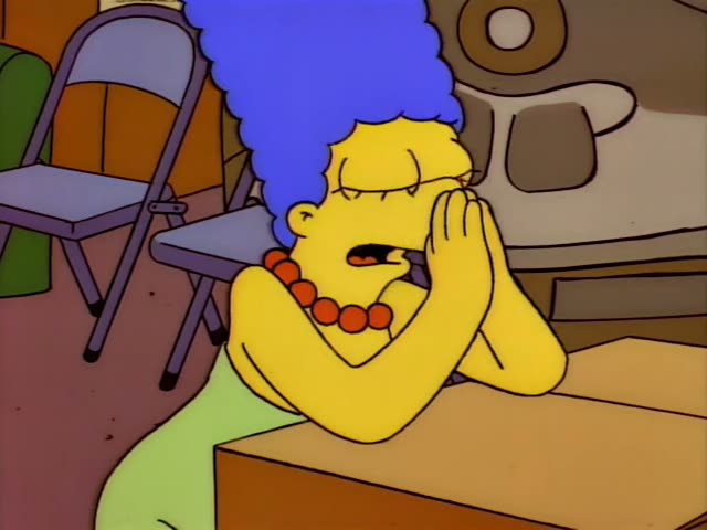 Marge Simpson sits at a wooden table in a room with folding chairs and a round grey-brown machine behind her