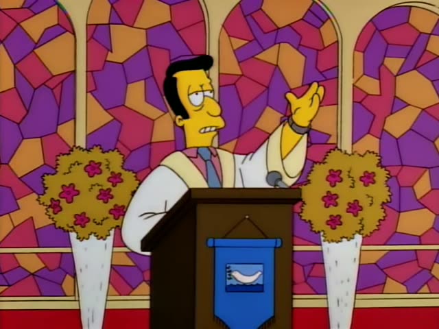 A thin man with black swept-back hair, a pink shirt, blue tie and cream vest stands behind a wooden podium with a microphone…