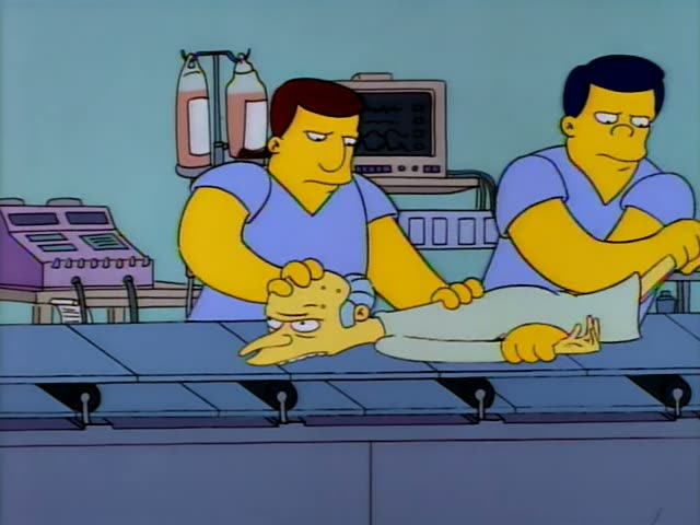 Mr. Burns lies on a long operating-room table while several men in blue scrubs work on him