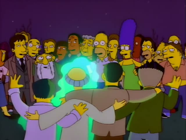 A crowd gathers outdoors under a dark purple night sky around a glowing green light and a small yellow figure…