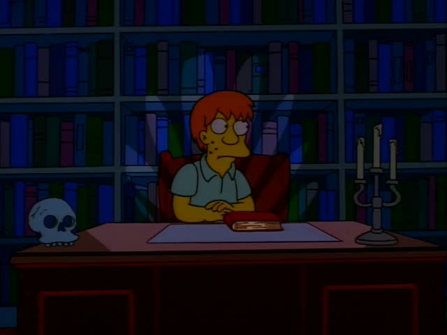 An unnamed man with short orange-red hair, round glasses and a light blue polo shirt sits behind a desk in a dim library…