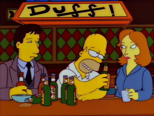 Homer sits at the bar in Moe's tavern beneath the orange Duff sign, next to a dark-haired man in a grey suit and tie…