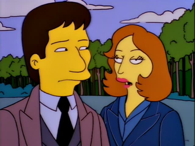 A slim, brown-haired man in a pinkish-brown coat, dark tie and grey vest stands beside a woman with shoulder-length…