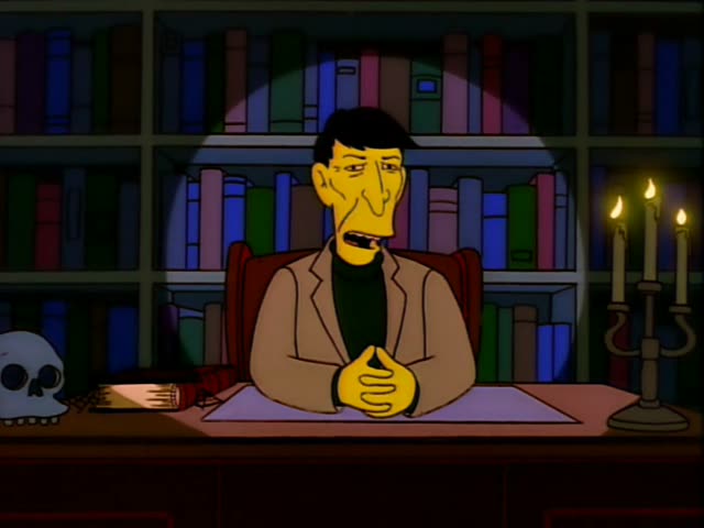 A thin, dark-haired man in a tan jacket over a dark green turtleneck sits in a red leather armchair behind a desk, hands…