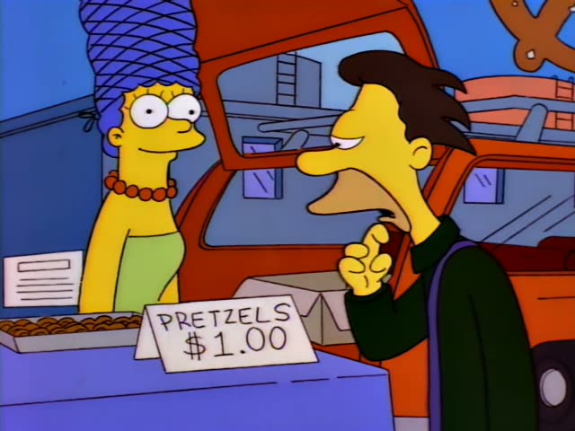 Marge Simpson stands behind a blue counter at a pretzel stand while a dark-haired man in a green jacket with a purple strap…