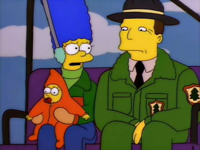 Marge Simpson sits on a purple chairlift seat holding baby Maggie in an orange hooded bunting with a pacifier, next…