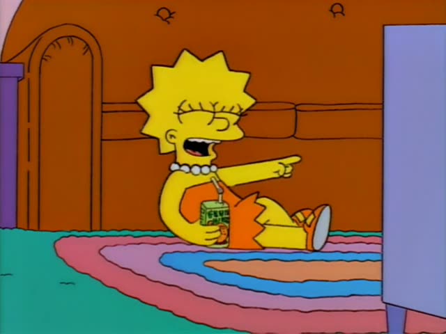 Lisa sits on a rainbow rug in the living room holding a green drink with a straw, pointing at the television and laughing…