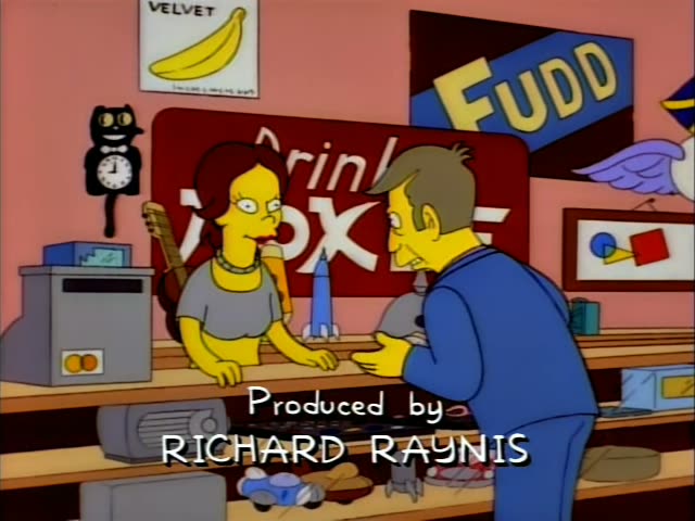 Troy McClure leans over a shop counter and gestures toward a woman with reddish-brown hair, glasses, a grey top and a beaded…