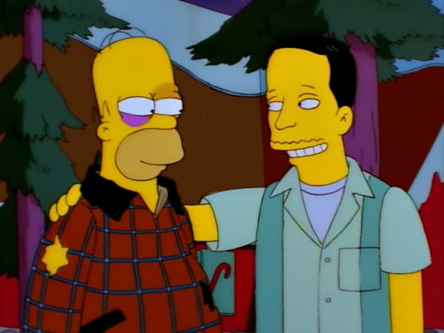 Homer Simpson stands outdoors beside a thin, dark-haired man in a light green vest over a pale shirt, with his arm draped…