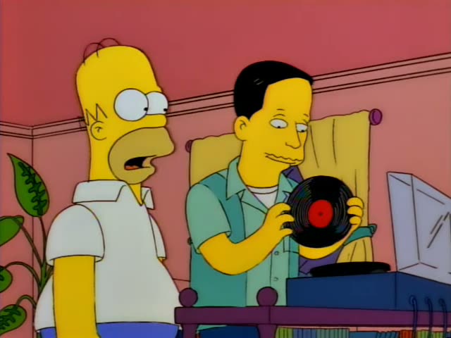 Homer Simpson stands beside a thin man with slicked black hair in a teal button-up shirt in a pink room with a potted plant…