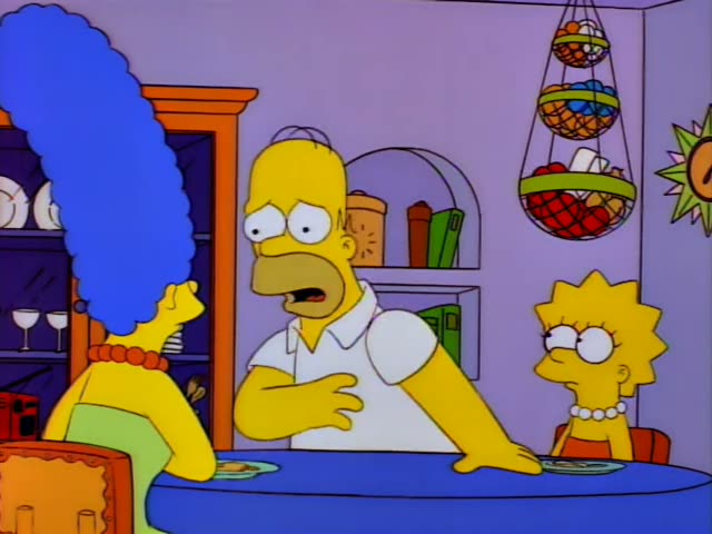Homer Simpson stands at the kitchen table beside Marge, whose tall blue beehive is in the left foreground, and Lisa sits…