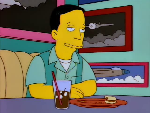 Marge Simpson with her tall blue beehive, Bart and Lisa Simpson sit in a diner booth beside a black-haired man in a light…
