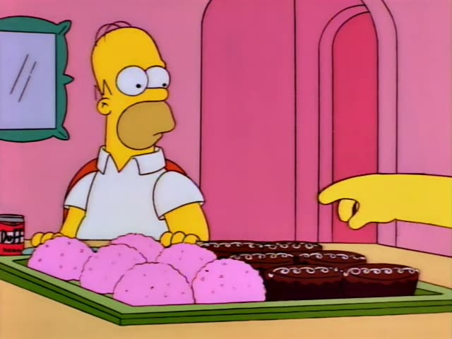 Homer Simpson stands at a counter behind a green tray of pink frosted cookies and chocolate brownies with white icing…