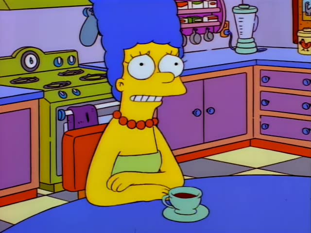 Marge sits at a kitchen table with her arms folded, facing Homer, who is bald and wears a white shirt, and a dark-haired man…