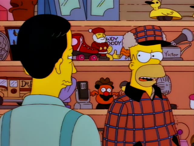 Homer stands at the right of a cluttered novelty shop facing a slim man with black hair, a light blue shirt and suspenders…