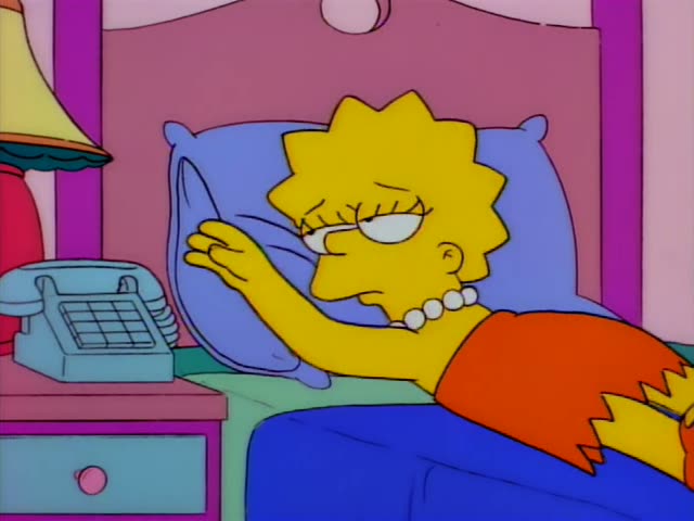 Lisa lies in bed under a blue blanket and reaches for a light blue pillow beside a bedside table holding a telephone…
