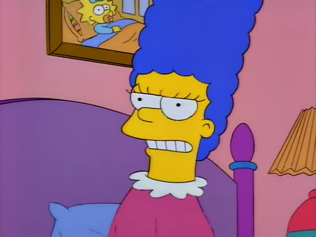 Marge sits up in a purple-headboard bed in a pink pink-walled bedroom, wearing a pink top with a white collar…