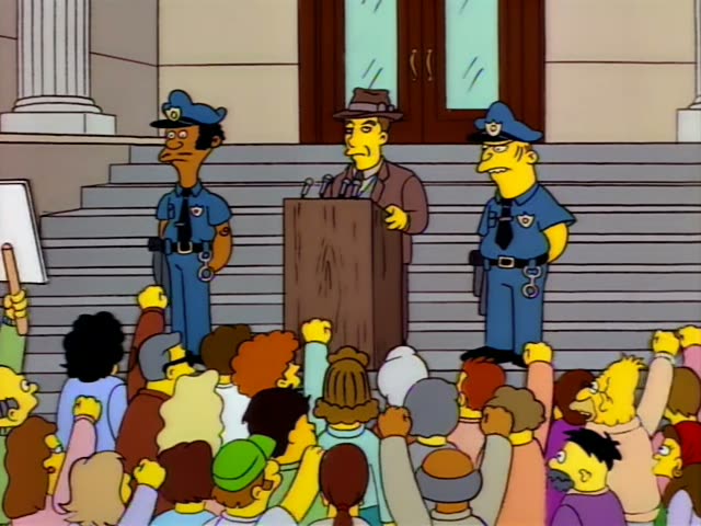 A man in a brown fedora and suit stands behind a wooden podium on courthouse steps, gesturing with one hand as he speaks…