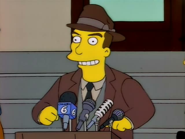 A slim, one-off man in a brown fedora and brown suit with a dark tie stands behind a podium crowded with microphones…