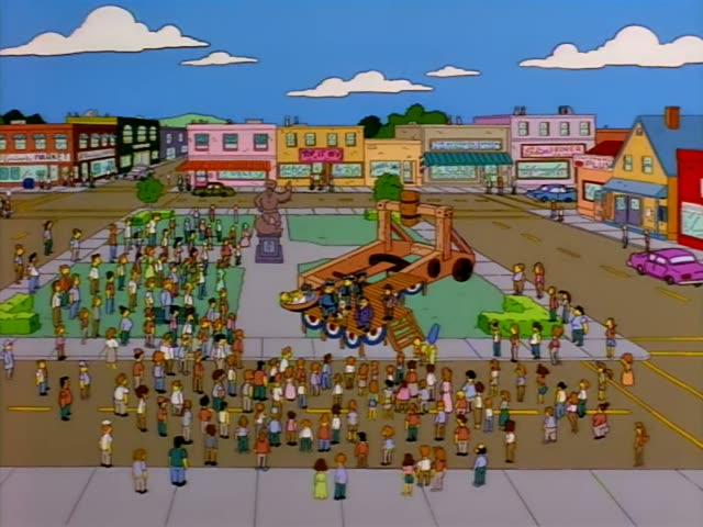 Homer Simpson lies strapped on his back in the bucket of a large wooden catapult on a platform in a town square…