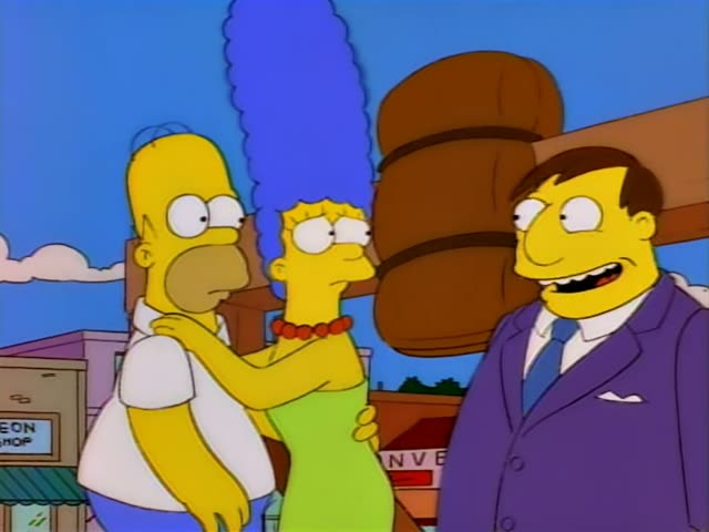 Homer and Marge stand with a man in a purple suit and blue tie in front of a large wooden barrel structure beside a street…