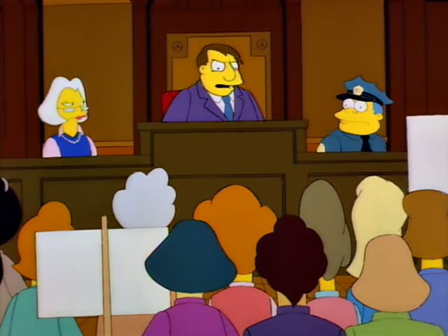 A heavyset man in a purple suit and blue tie sits at a raised judge's bench, with a white-haired woman in a blue dress…