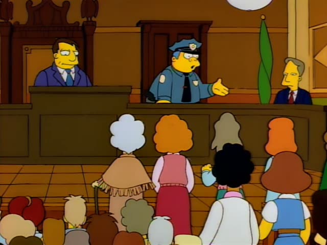 Police Chief Wiggum stands behind a courtroom bench and gestures with open hands, then points and sweeps one arm out…