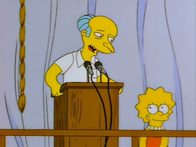 Mr. Burns stands at a wooden podium with microphones and talks while raising a finger, then grips a long golden rope hanging…