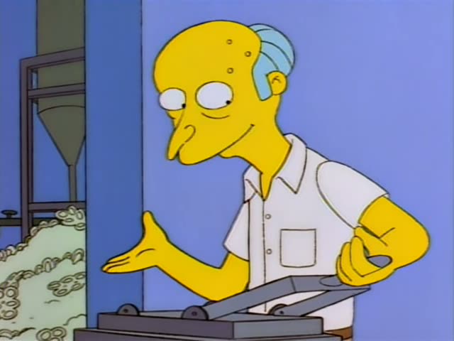 Mr. Burns stands in a white shirt beside a machine, gesturing with his hand and then pointing at a pile of interlocked…