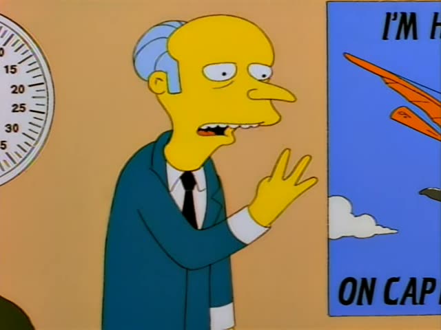 Mr. Burns stands in a office-like room in a dark teal suit and black tie, gesturing and speaking with his hands